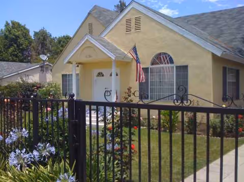 Yellow single-story senior living exterior with front porch, American flag, and black wrought-iron fence.