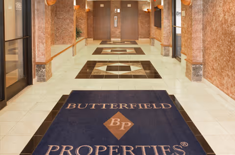 Hallway with tiled floors, elevator doors, and a Butterfield Properties mat in a senior living facility.