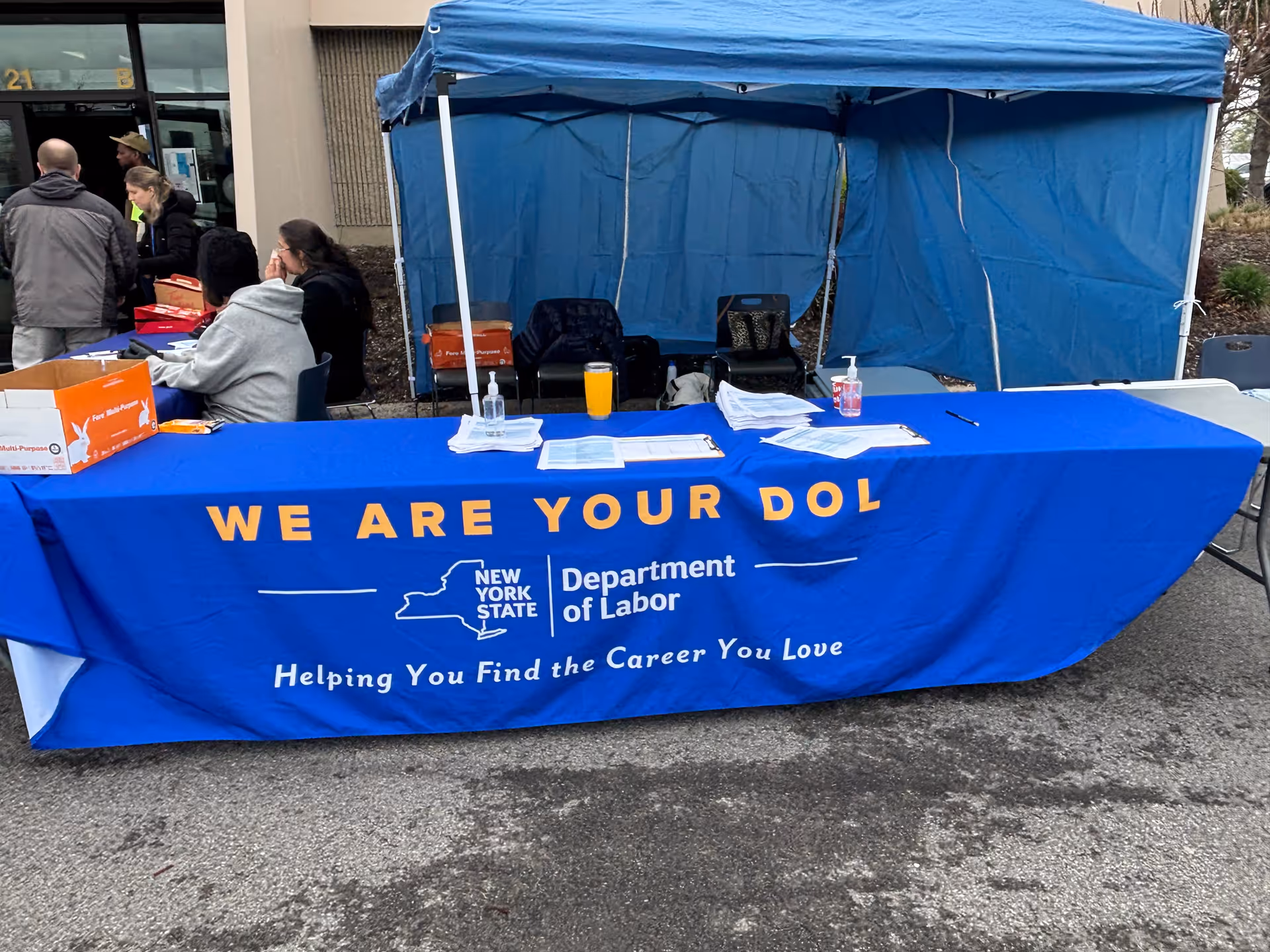 Outdoor information booth with a blue tablecloth, tent, and people nearby.