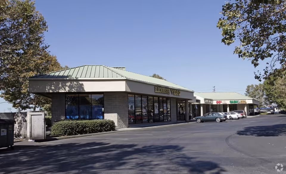 Exterior view of a strip mall storefronts, including Blockbuster Video and parked cars.