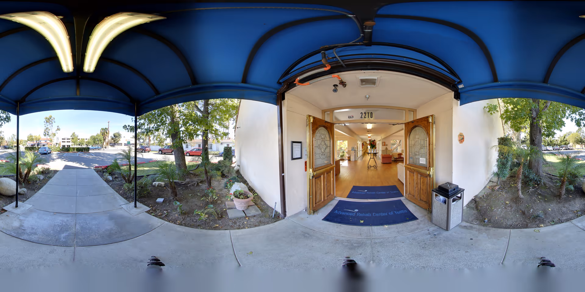 Covered entrance with a blue awning and open double wooden doors leading into a senior care facility, with a walkway and landscaping.
