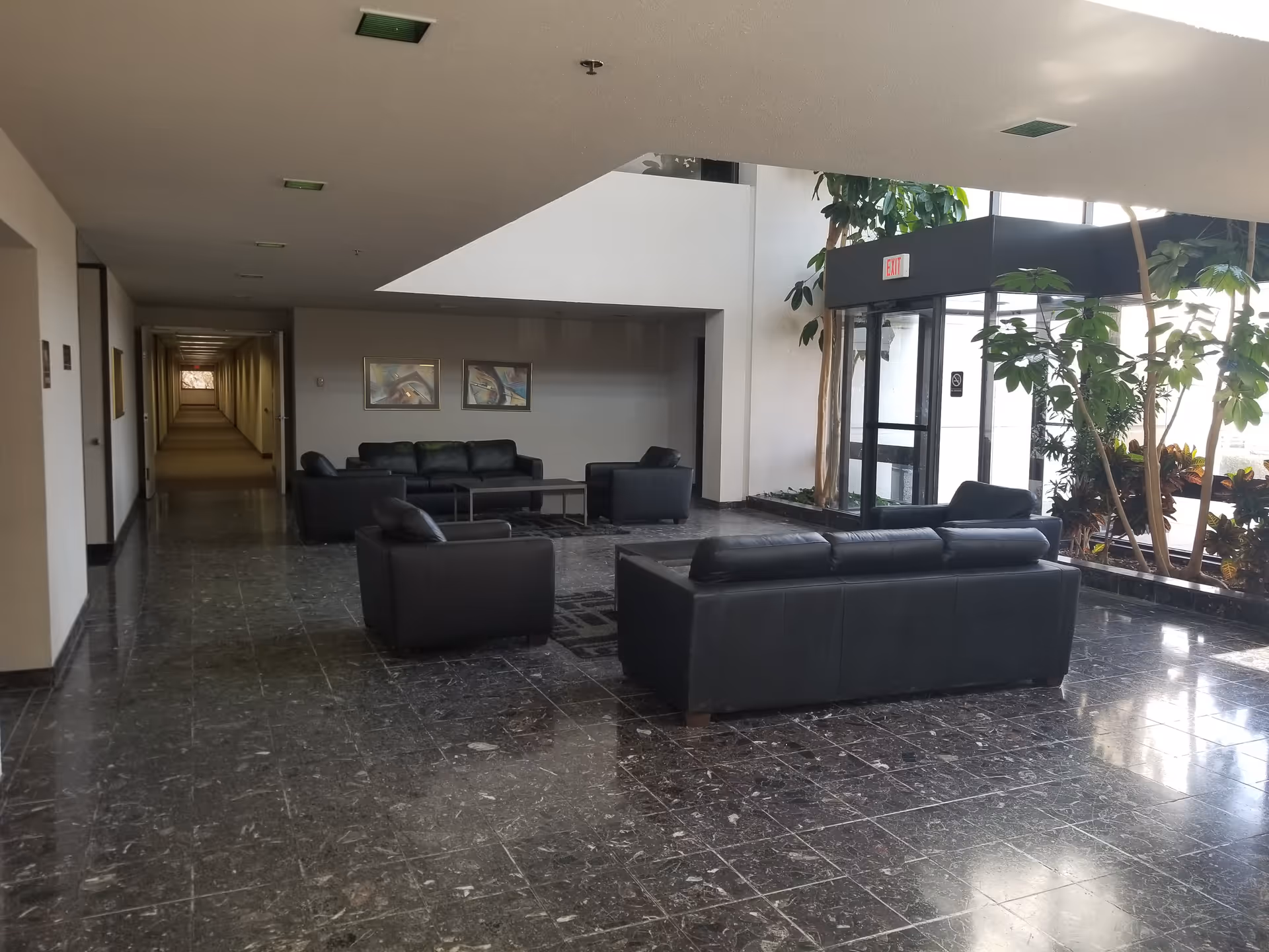 Spacious lobby area with black leather sofas, tiled floor, and indoor plants near glass entry.