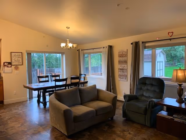 A cozy living and dining area with a small gray couch and a green recliner chair next to a wooden side table with a lamp. The dining area has a wooden table with six chairs under a chandelier light fixture. Large windows with curtains provide natural light and a view of a fenced backyard with trees and a shed.