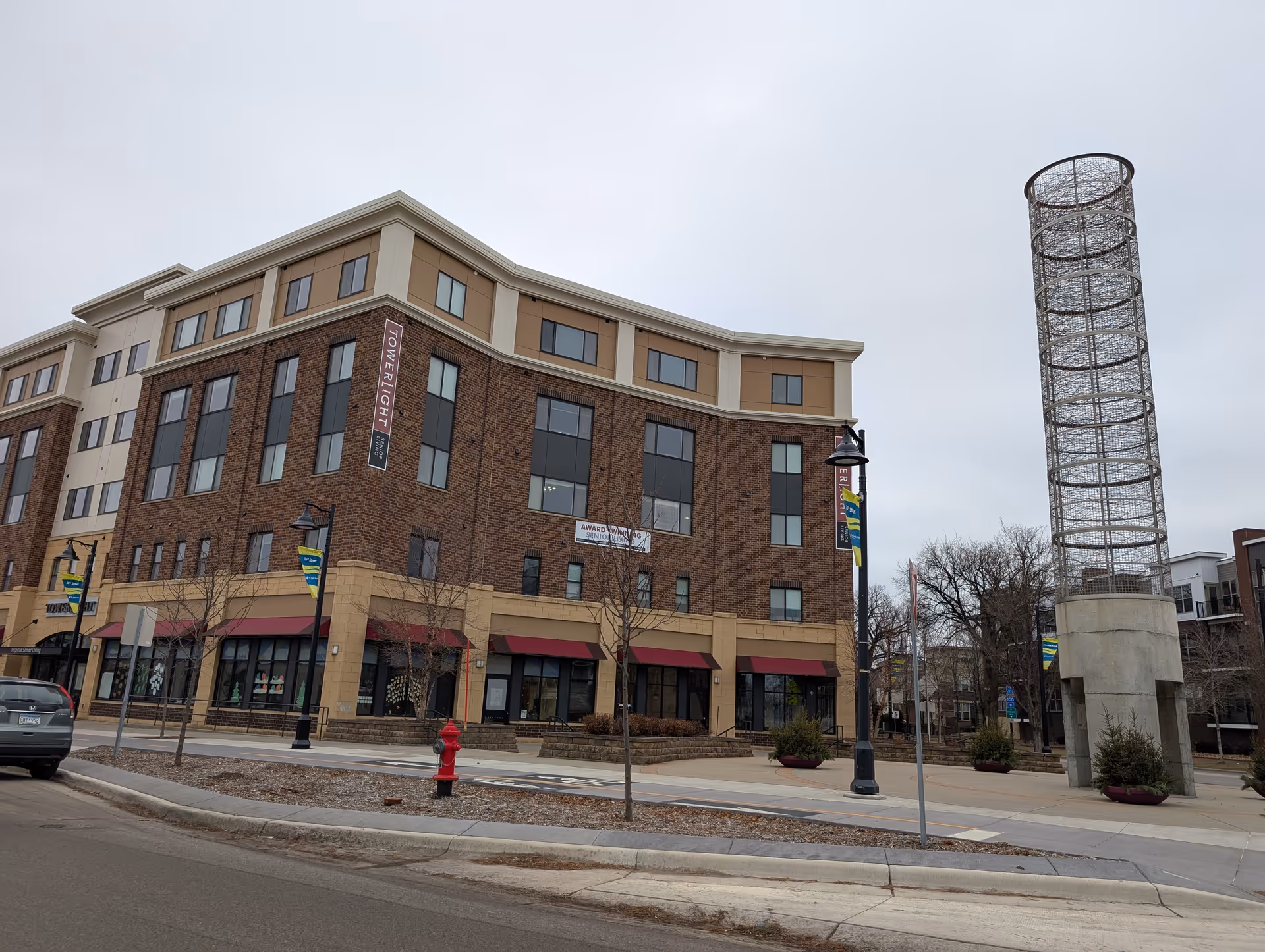 Brick and tan four-story building with storefronts, a vertical 'Towerlight' sign, and a tall wire-frame tower sculpture by the sidewalk.