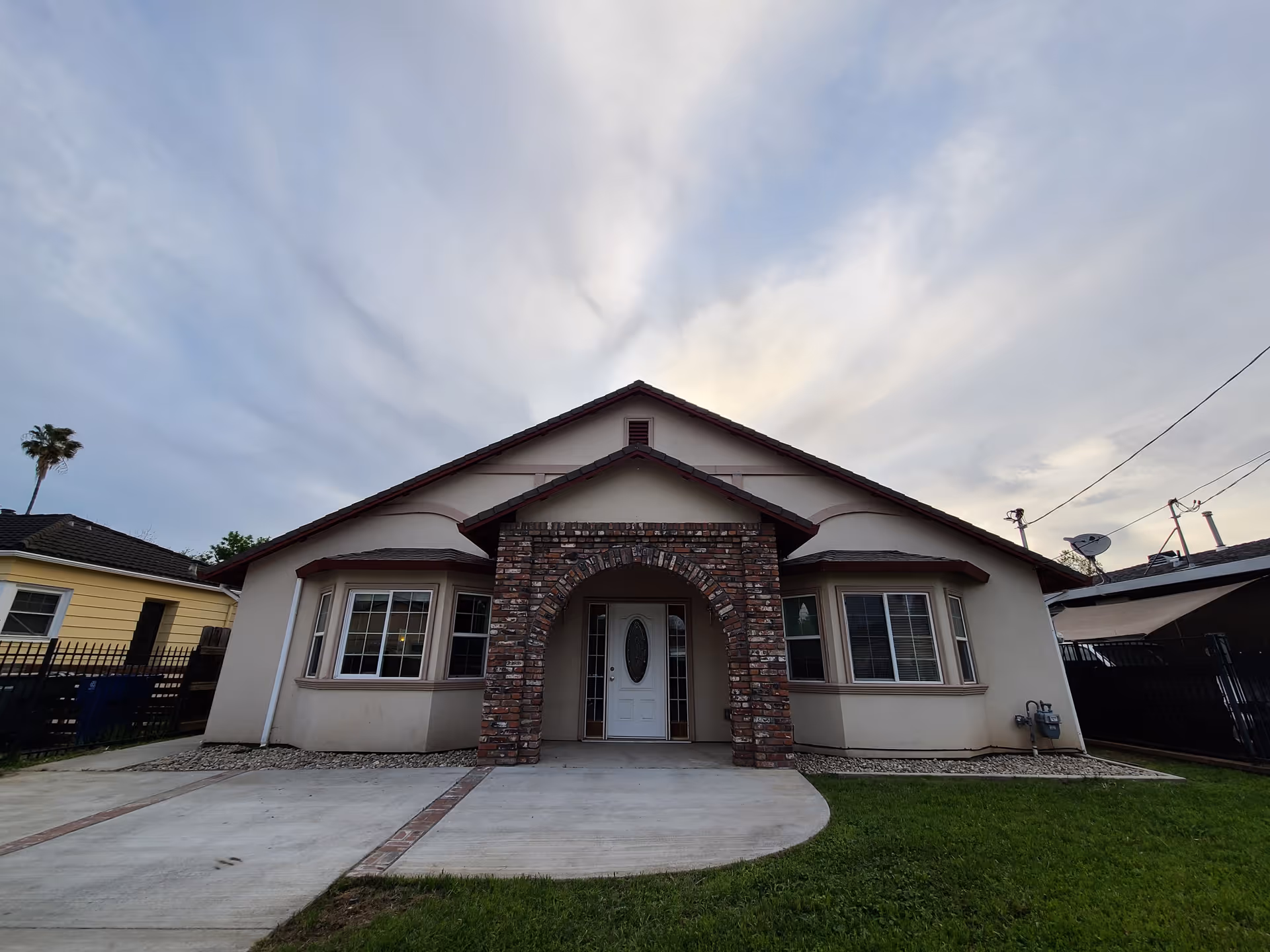 Front exterior of a single-story house with a brick arched entry, driveway and small lawn under a cloudy sky.