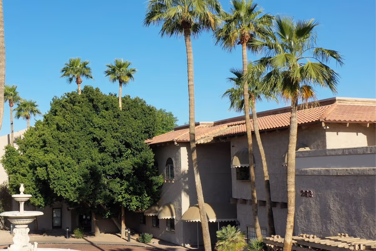 Exterior view of a senior living facility building with beige stucco walls and a red tile roof, surrounded by tall palm trees and a large leafy tree. A white stone fountain is visible in the foreground under a clear blue sky.