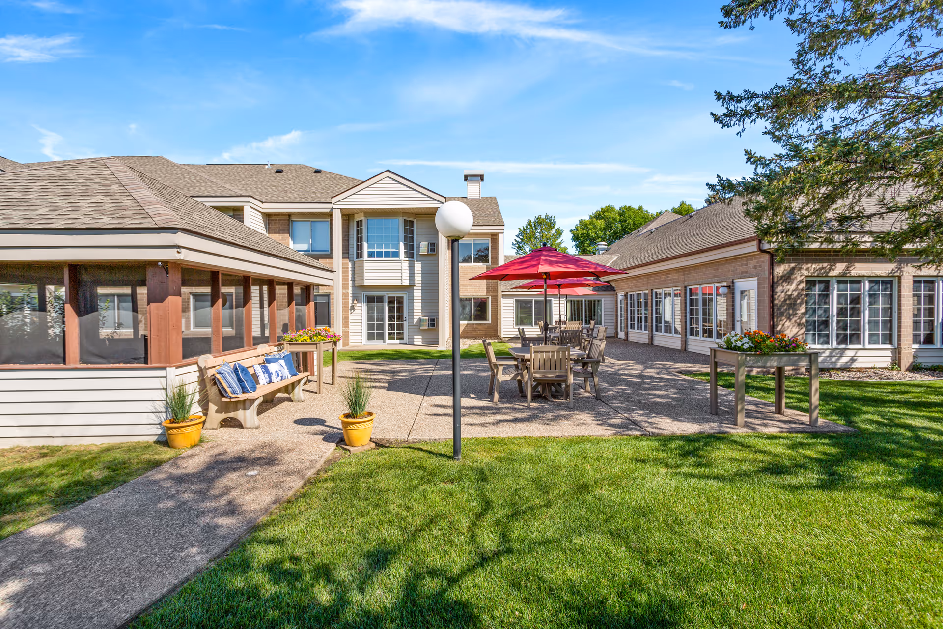 Outdoor patio area at Riverwood Village with tables and chairs under red umbrellas, a bench with blue and white cushions, potted plants, and a well-maintained lawn surrounded by buildings under a clear blue sky.