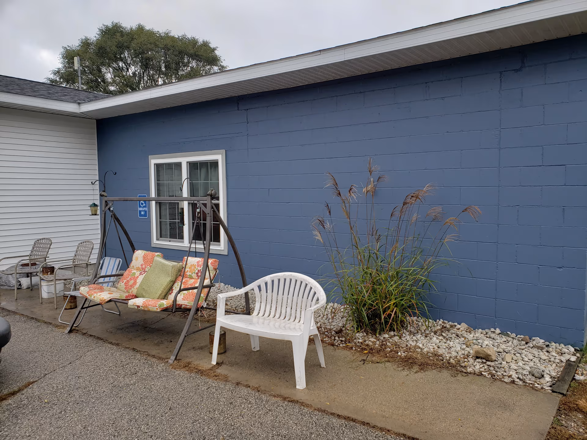 Outdoor seating area beside a blue-painted cinderblock building wall with a window, a swinging bench, a plastic chair, and ornamental grasses.