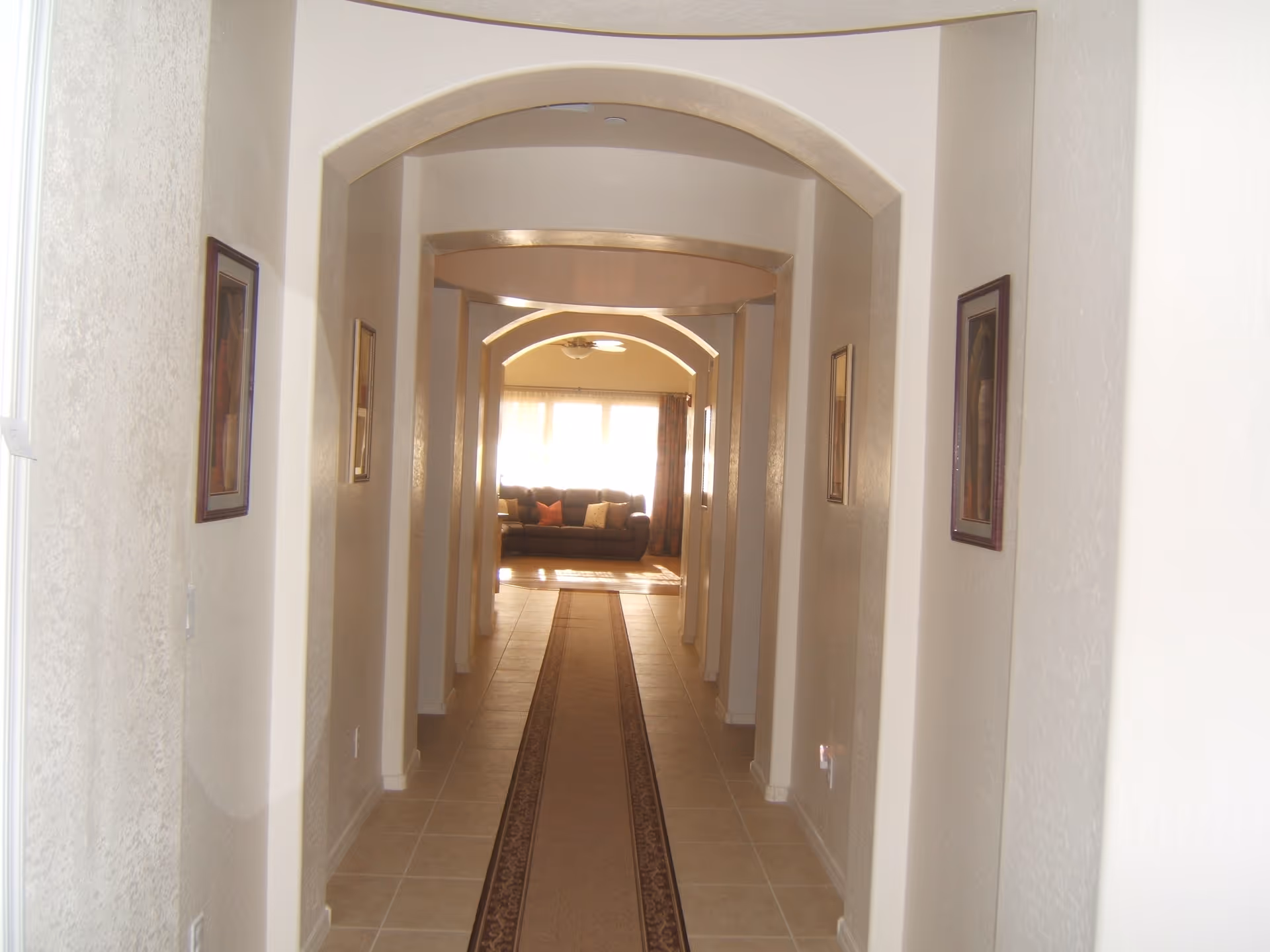 Long tiled interior hallway with arched doorways leading to a sunlit living room with a sofa at the end.