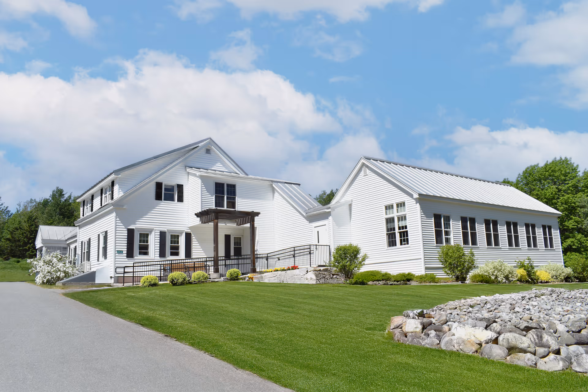 White multi-wing building with a covered entrance, manicured green lawn and stone landscaping under a blue sky.