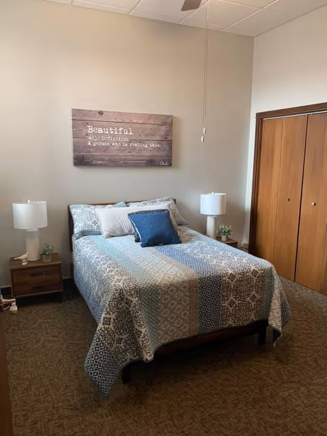 A neatly made bed with blue patterned bedding flanked by nightstands and lamps in a simple bedroom with wooden closet doors and wall art above.