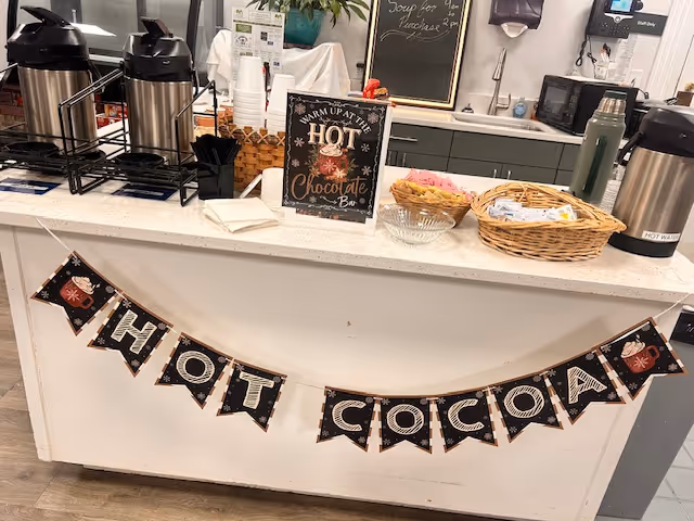 Service counter with hot chocolate sign, cups, and beverage dispensers inside a facility.