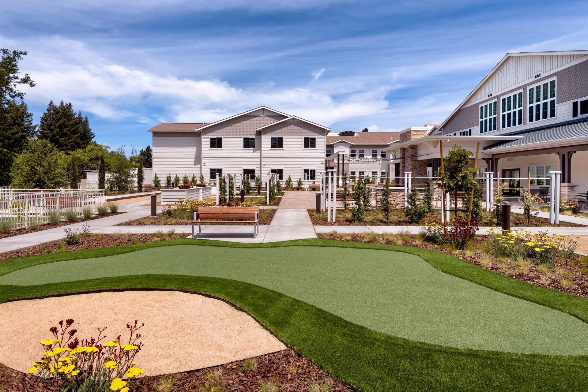 Outdoor garden area at Clearwater at Sonoma Hills featuring a putting green, landscaped flower beds with yellow flowers, a wooden bench, paved walkways, and a white multi-story building in the background under a partly cloudy blue sky.