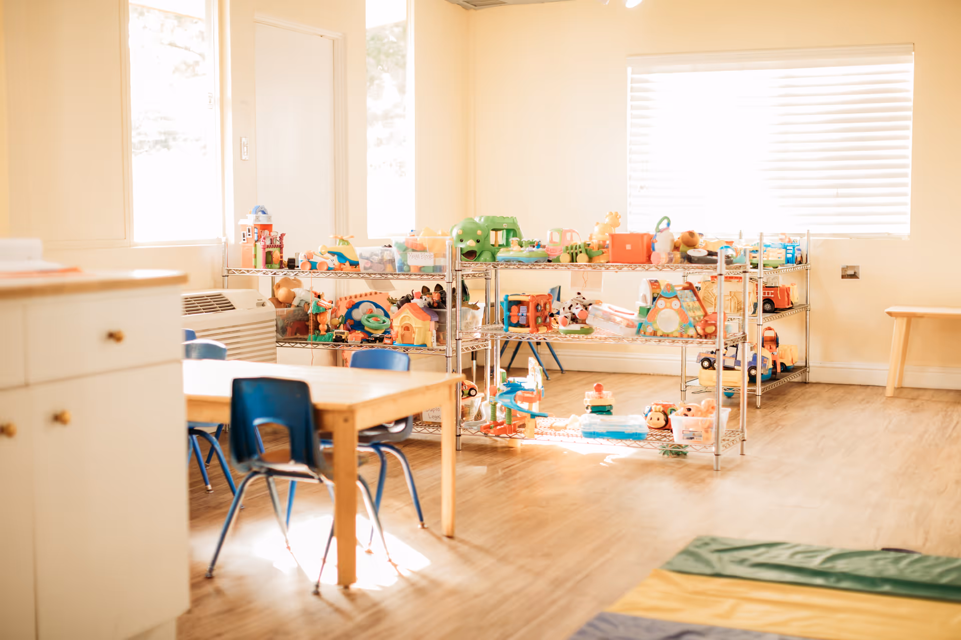 Bright playroom with shelves of toys, small tables, and blue chairs.