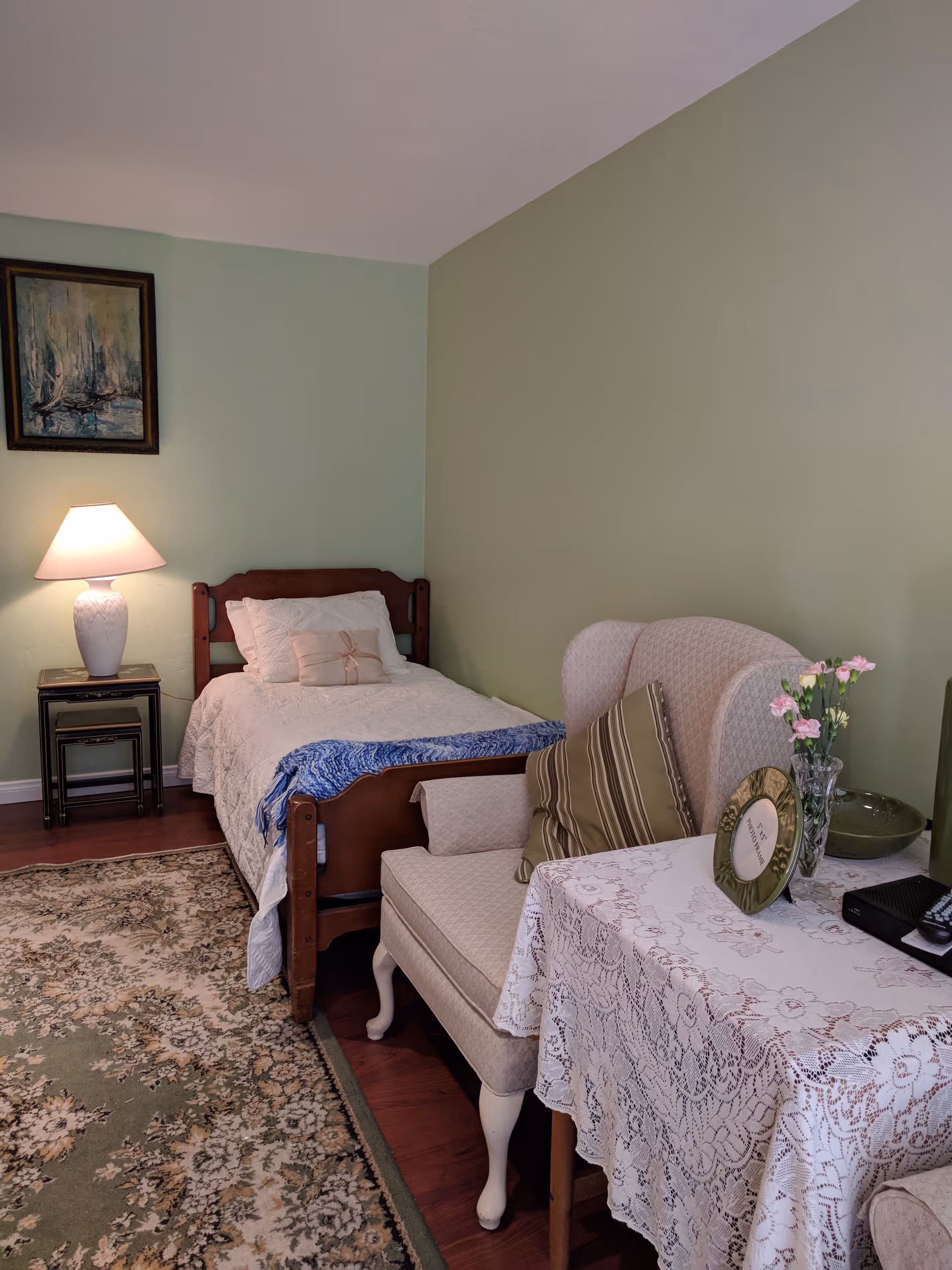 A cozy bedroom with a single wooden bed covered with white bedding and a blue throw blanket. Next to the bed is a small side table with a white lamp. There is a patterned area rug on the wooden floor. A cushioned armchair with a striped pillow sits beside a table covered with a lace tablecloth, holding a vase with pink flowers and a photo frame. The walls are painted in light green and beige tones, and a framed painting hangs above the lamp.