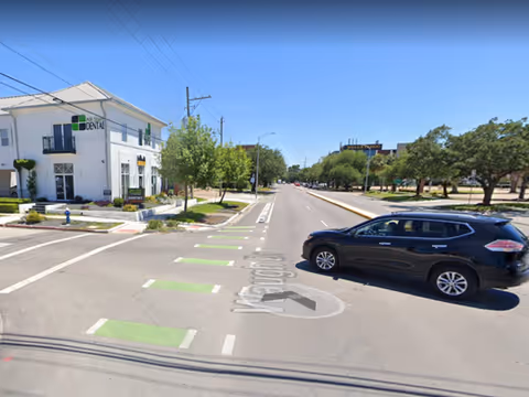 Wide street intersection with a white two-story building, trees, and a dark SUV crossing.