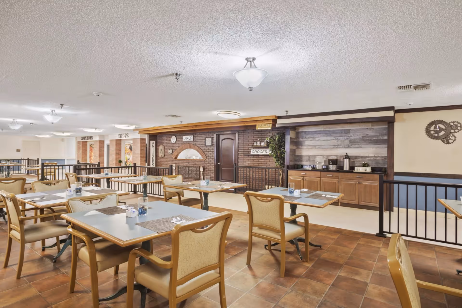 A dining area with multiple tables and chairs arranged neatly. The tables have placemats, utensils, and small decorative items. The background features a brick wall with signs reading 'DINER,' 'GROCERIES,' 'ANTIQUES,' and 'DOWNTOWN SHOPS,' giving the space a quaint, small-town feel. There is a counter area with a coffee and water station on the right side.