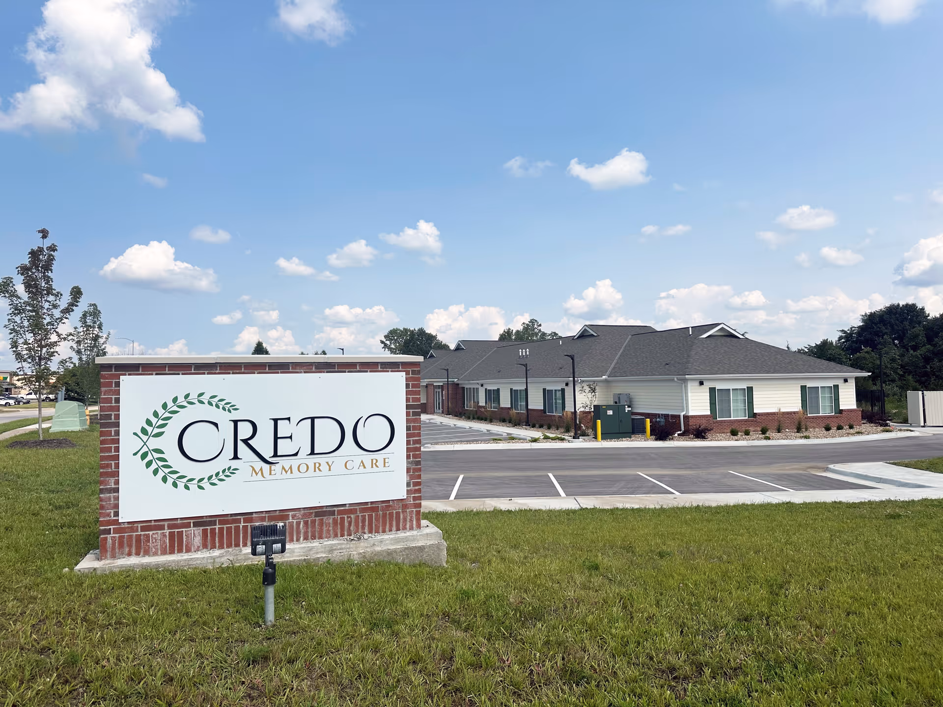 Exterior view of Credo Memory Care - Laurel Residence building with a large sign in the foreground displaying the facility's name. The building is single-story with white siding and brick accents, surrounded by a parking lot and green grass under a partly cloudy sky.