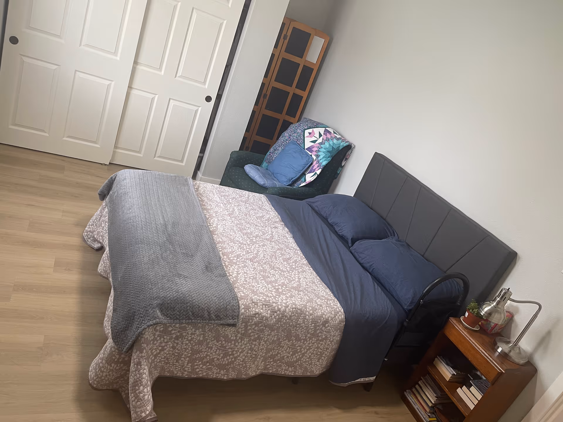 A neatly made bedroom with a bed, nightstand, lamp, and chair placed by the wall.