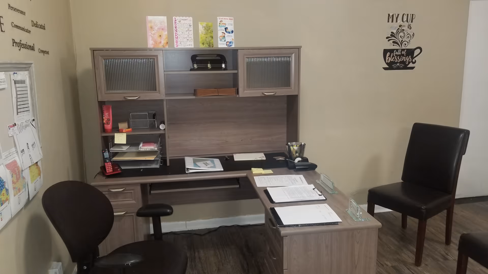 A compact office desk with paperwork, shelves, and two chairs in a neutral room.