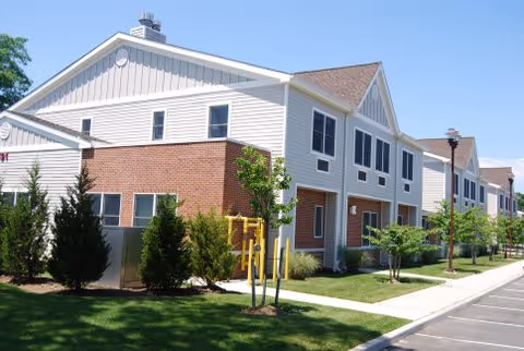 A two-story senior living building with brick lower facade, beige siding, trees, and a sidewalk.