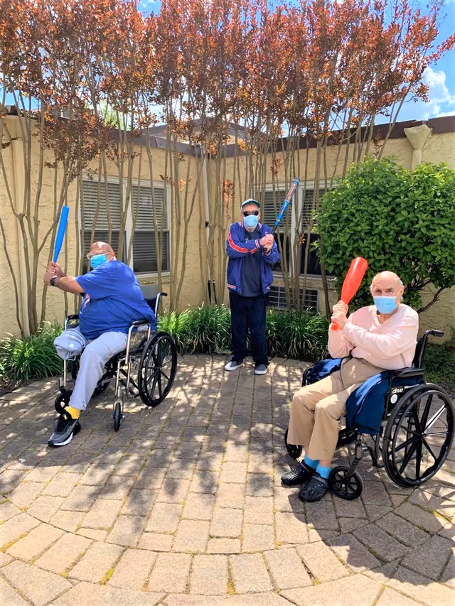Three elderly men wearing face masks playing with plastic baseball bats in an outdoor courtyard area. Two men are seated in wheelchairs holding bats, and one man is standing in the middle also holding a bat. The courtyard is paved with bricks and surrounded by plants and trees with a building wall in the background.