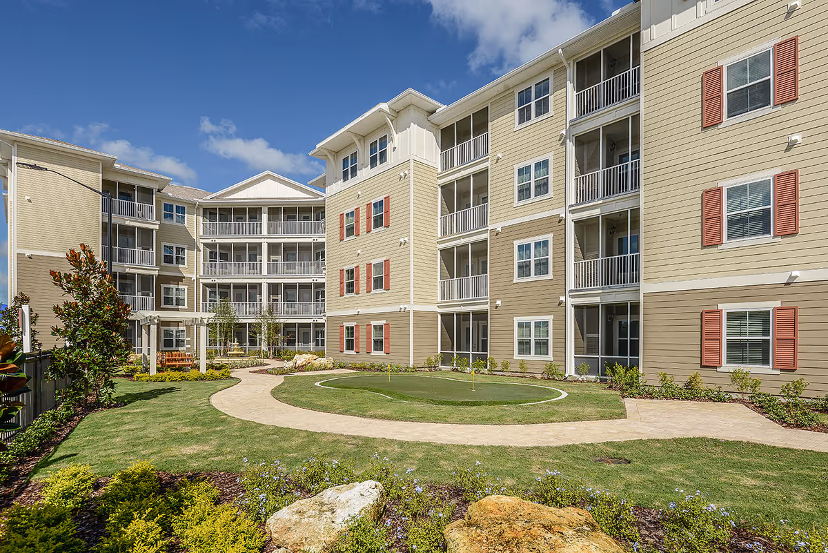 Exterior view of a multi-story beige apartment building with red shutters and screened balconies surrounding a landscaped courtyard with a putting green, walking path, and garden beds under a blue sky with some clouds.
