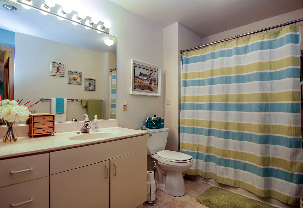 Bathroom in a senior living facility with sink, mirror, toilet, and striped curtain.