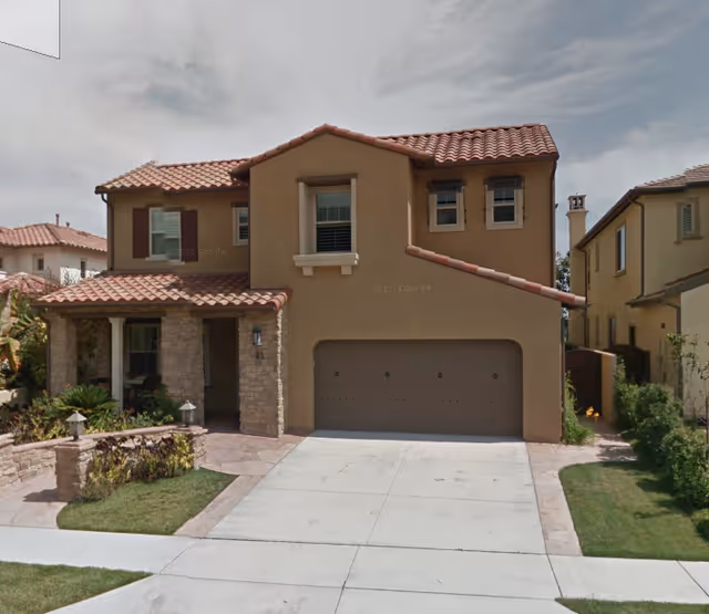 Two-story tan stucco house with red-tiled roof, attached garage, and small front yard.