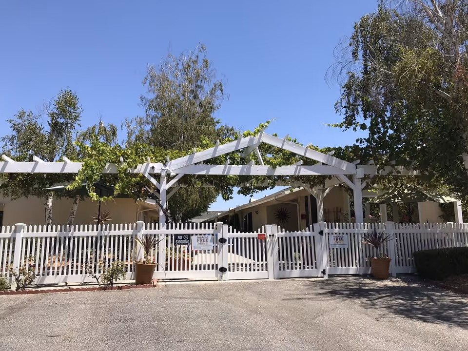 White gated entrance with a pergola, trees, and potted plants at a senior living facility.