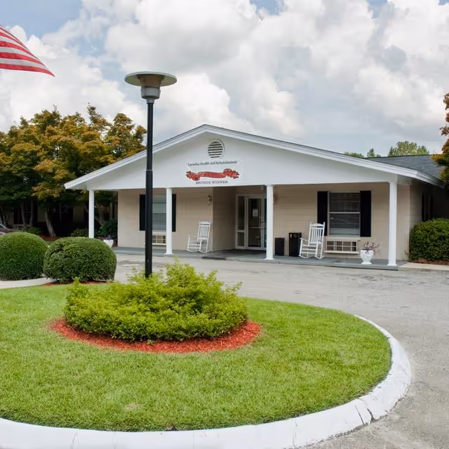 Exterior view of Camellia Health & Rehabilitation building with a covered entrance, two white rocking chairs on the porch, manicured bushes and grass in front, and an American flag on the left side.