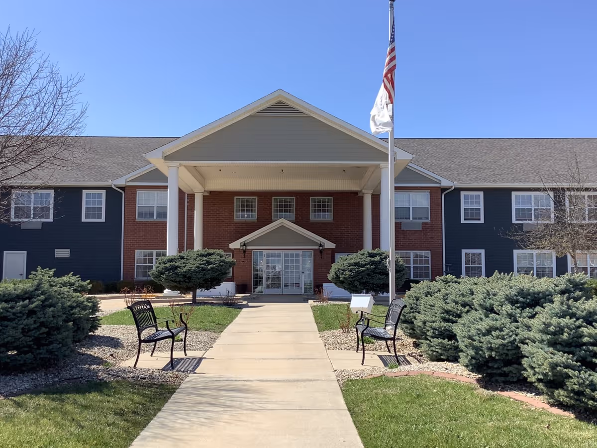 Front entrance of a two-story senior living building with a covered portico, flagpole, benches, and landscaped shrubs.