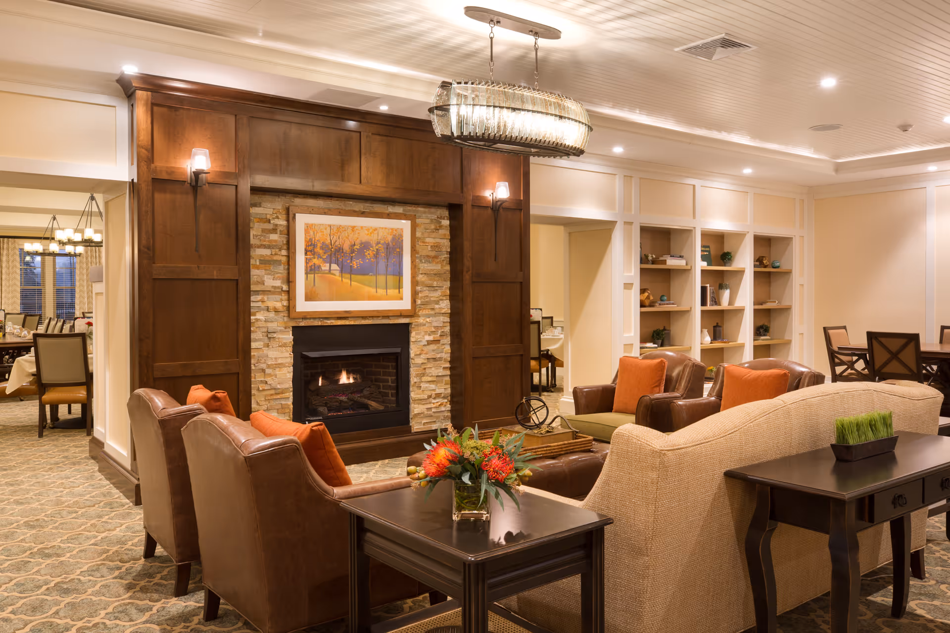 A cozy living room area in The Residence at Orchard Grove featuring a stone fireplace with a wooden mantel, a framed painting above it, and several comfortable chairs and sofas with orange cushions arranged around a coffee table. There are built-in shelves with decorative items on the right wall and a dining area visible in the background.