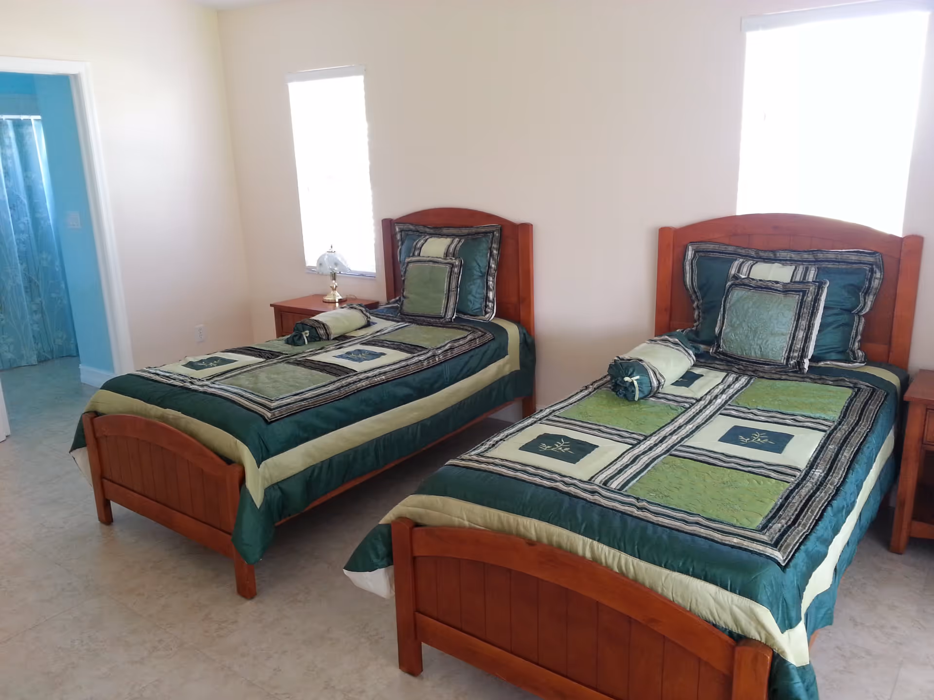 A bedroom with two single wooden beds, each made with green and beige patterned bedding and matching pillows. There are two windows behind the beds letting in natural light. A small wooden nightstand with a lamp is placed between the beds. In the background, a doorway leads to a bathroom with a blue shower curtain.