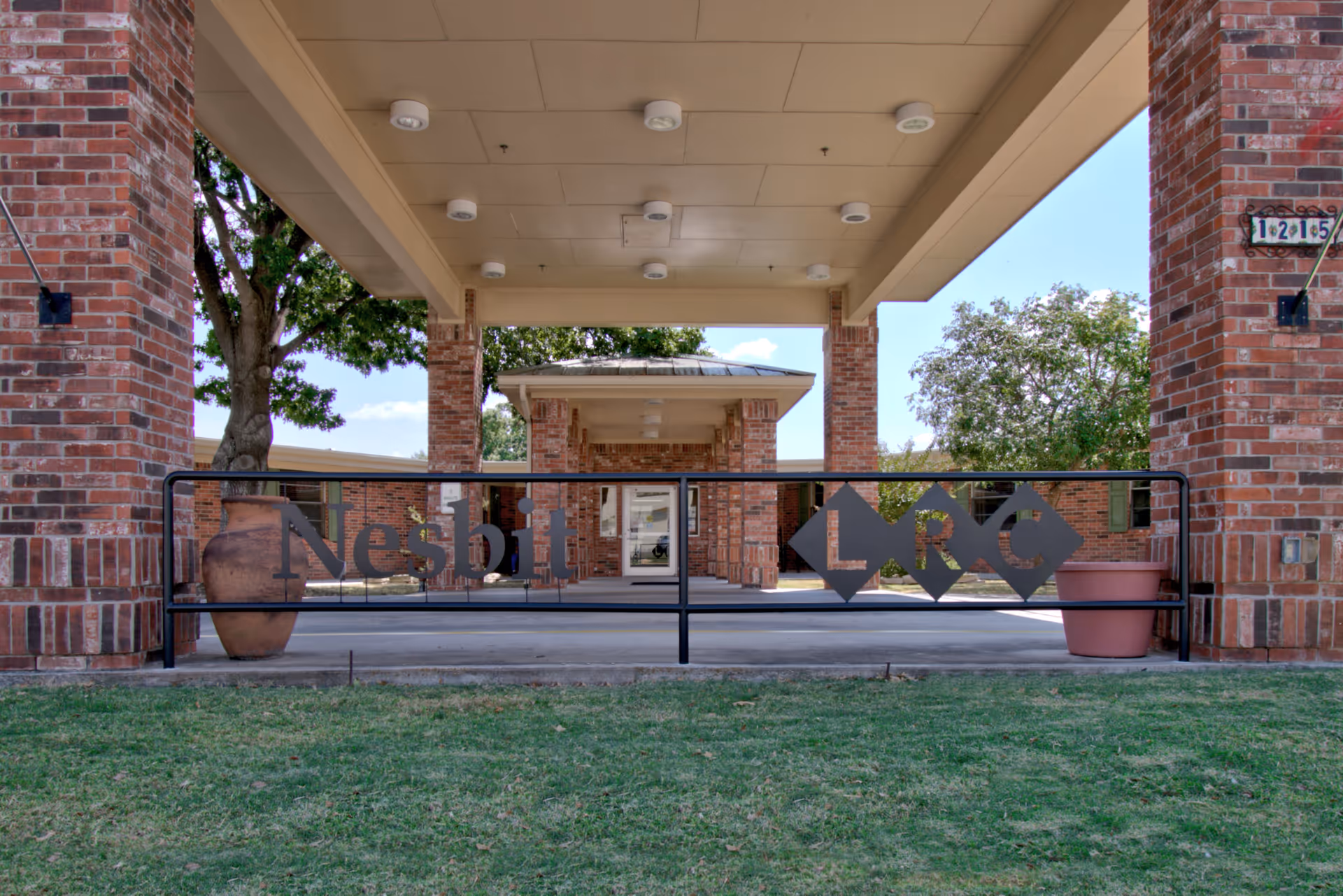 Entrance of Nesbit Living & Recovery Center showing a covered driveway supported by brick pillars, with a black metal gate featuring the name 'Nesbit LRC'. There are large potted plants on either side of the gate and trees in the background under a clear sky.