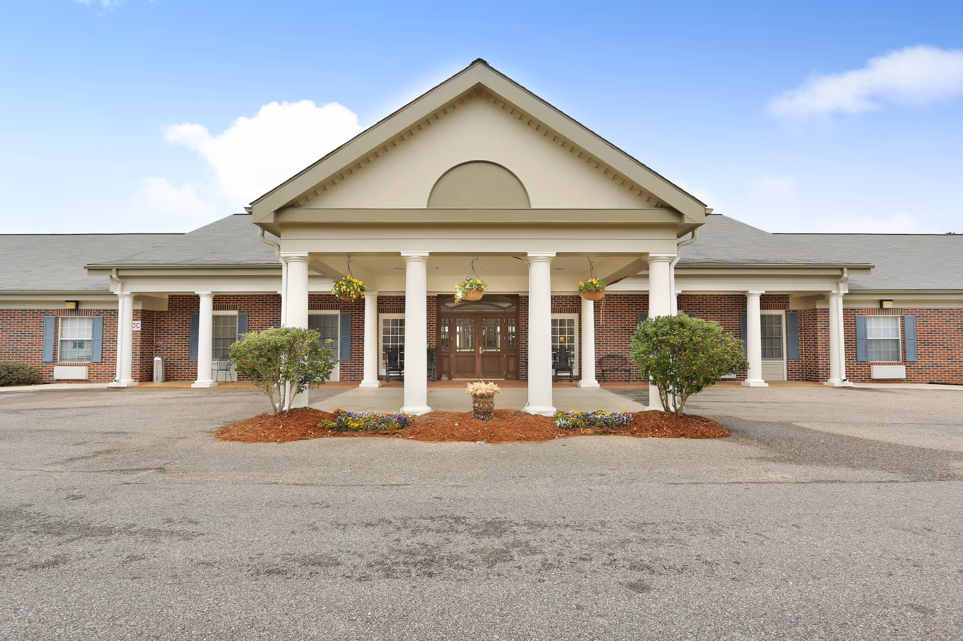Front entrance of a brick senior living building with white columns, hanging flower baskets, shrubs, and a paved driveway.