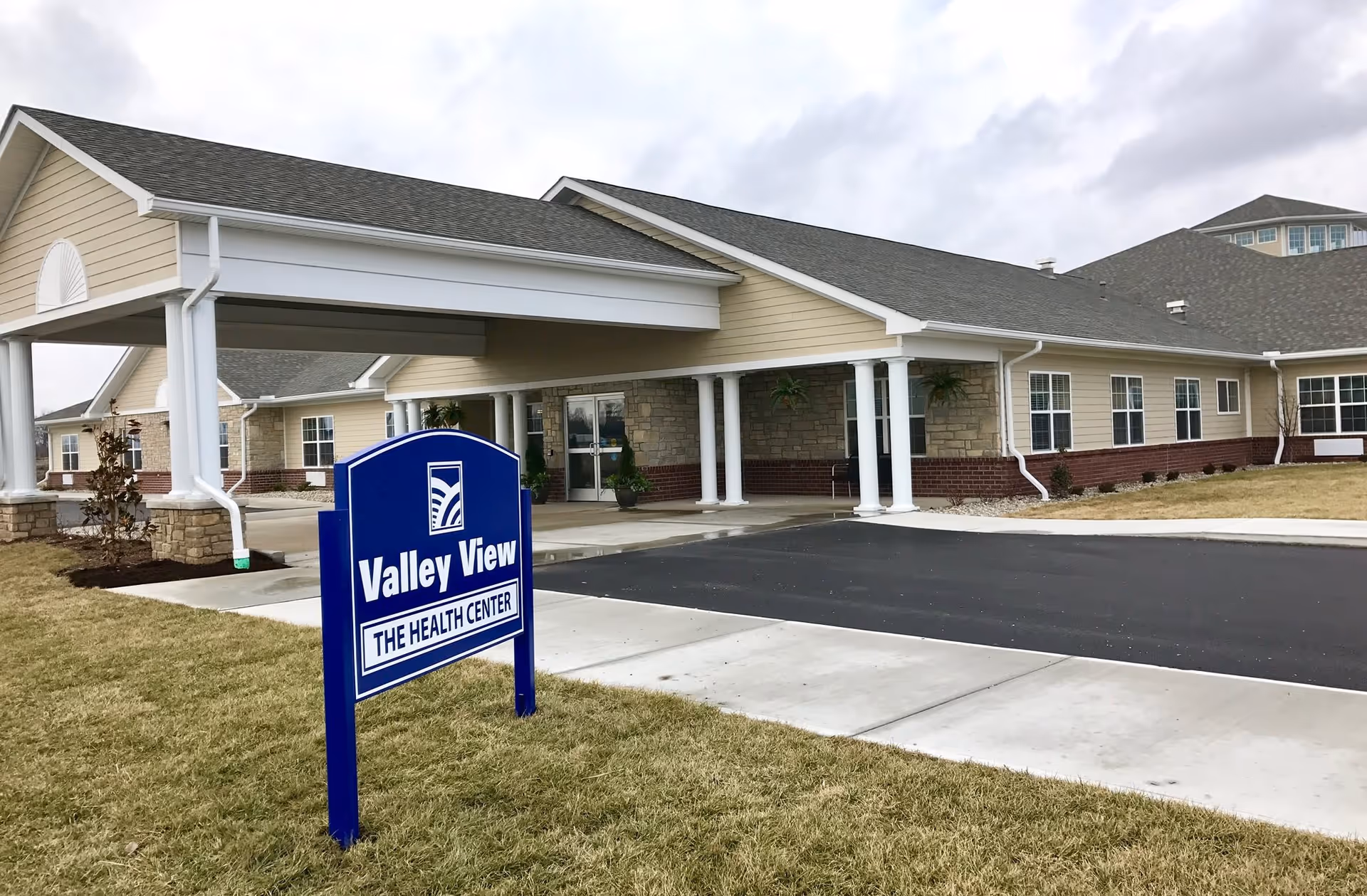 Exterior view of Valley View Health Center building with a covered entrance supported by white columns, beige siding, stone and brick facade, and a blue sign in the foreground on the lawn.