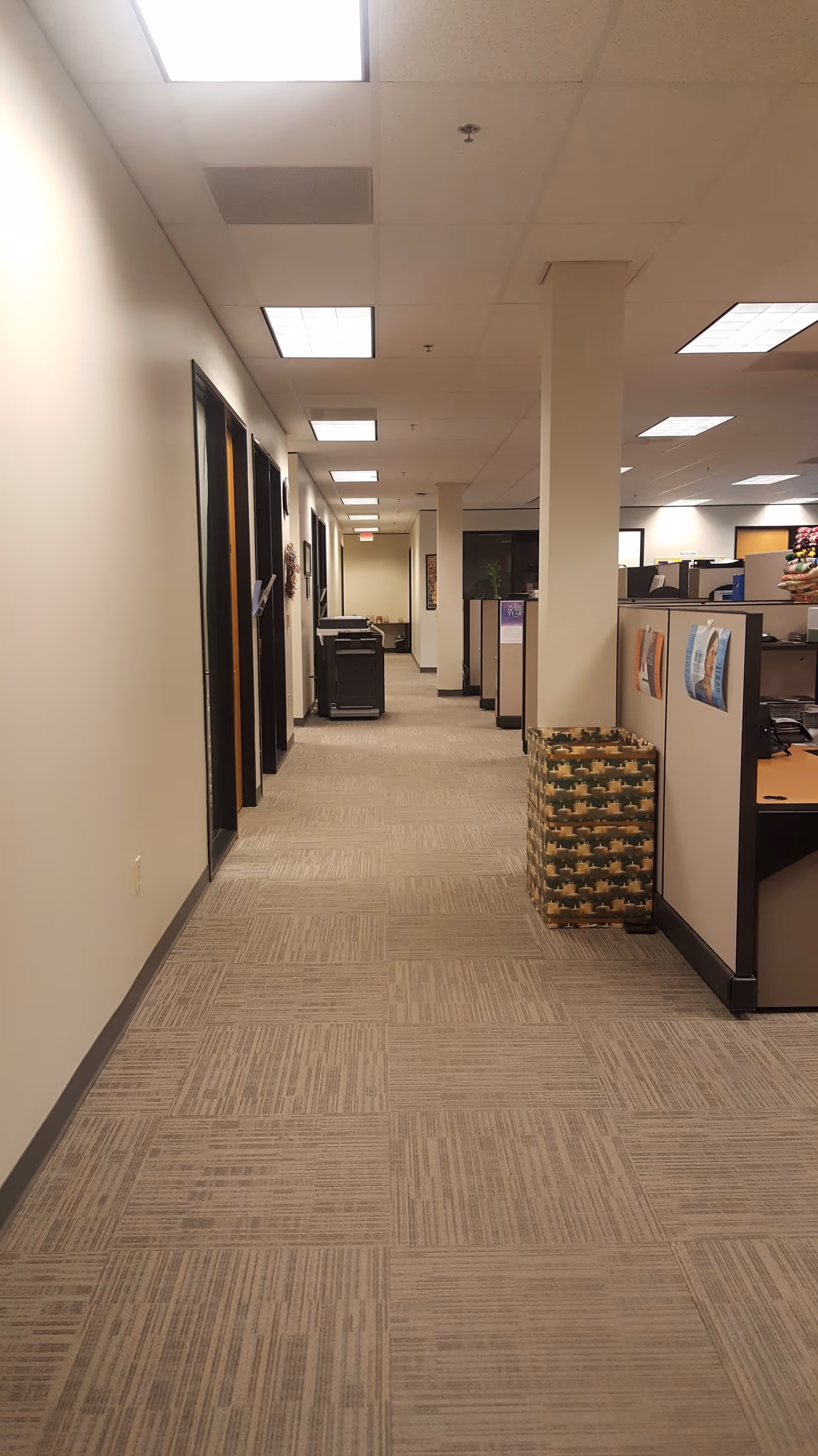 A long office hallway with cubicles, tiled carpet, and overhead fluorescent lighting.