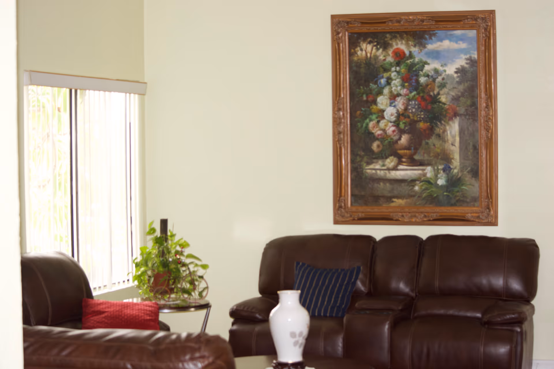 A cozy living room with dark brown leather sofas, a small round table with a green potted plant, a white vase on a coffee table, and a framed painting of a colorful flower bouquet hanging on a light-colored wall.