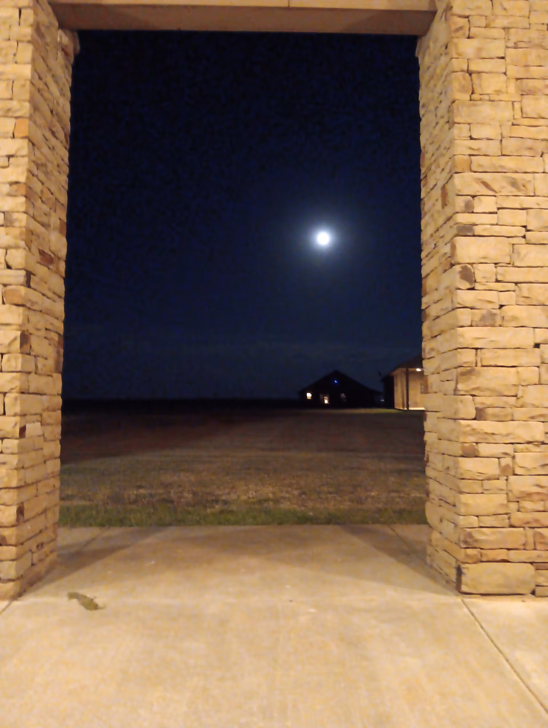 Night view through stone entrance pillars toward a distant lit building under moonlight.