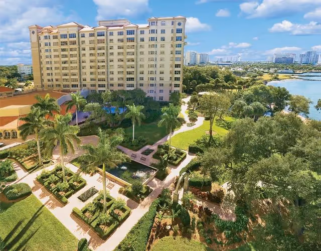 A large multi-story residential building surrounded by well-maintained gardens with palm trees, walking paths, and green lawns. The scene includes a waterfront view with a body of water and distant buildings under a partly cloudy blue sky.