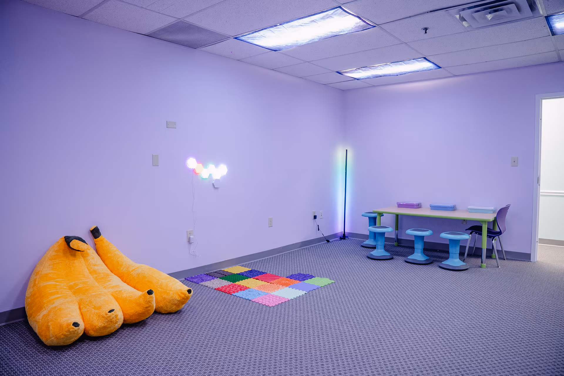 Lavender therapy room with plush orange cushions, a colorful rug, and child-sized seating.