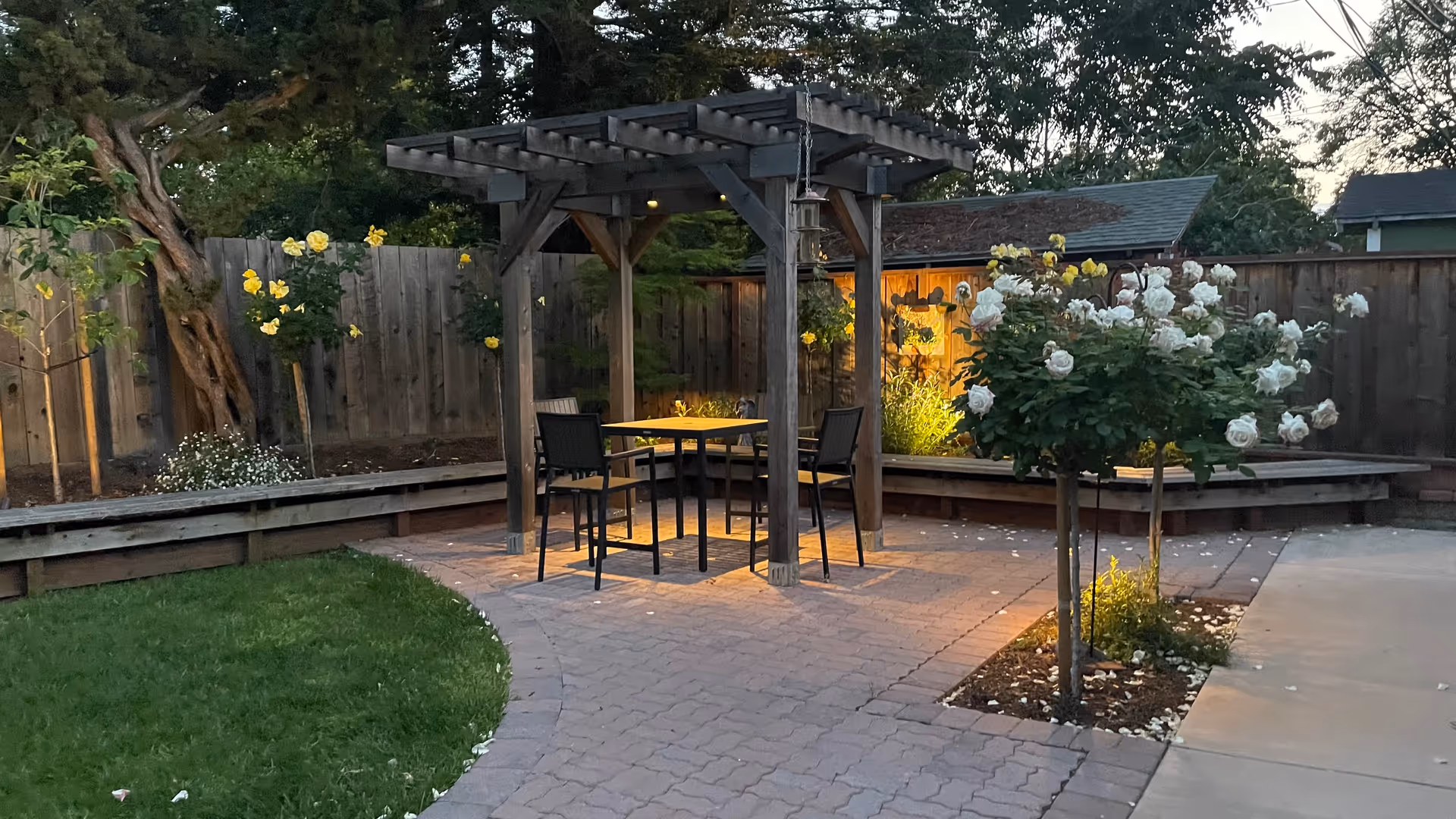 Outdoor patio under a wooden pergola with seating, rose bushes, and warm garden lighting.
