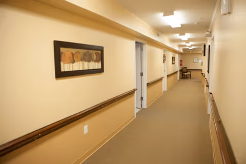 Long beige interior hallway with handrails, wall art, and doors leading to rooms in a senior living facility.