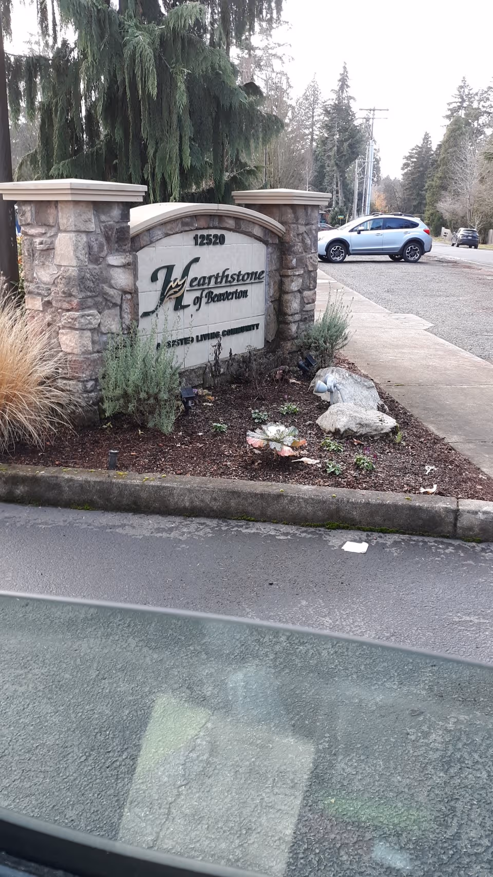 Stone entrance sign reading Hearthstone of Beaverton with plants and a parked car.