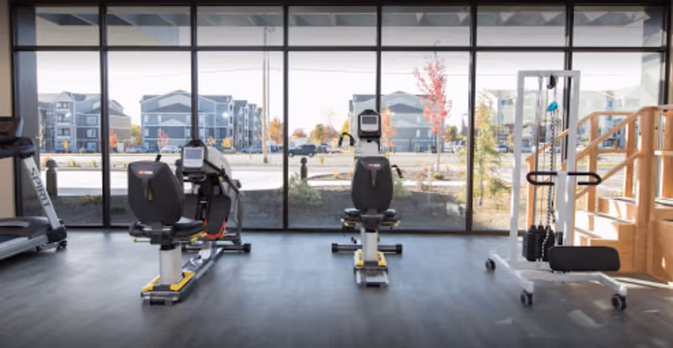 A fitness room with exercise equipment including two recumbent bikes, a treadmill, and a weight machine. Large floor-to-ceiling windows provide a view of an outdoor area with buildings and trees.