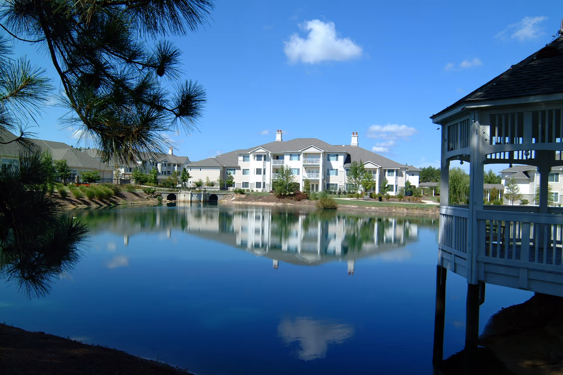 White multi-story senior living buildings and landscaping reflected in a calm pond with a gazebo at the right.