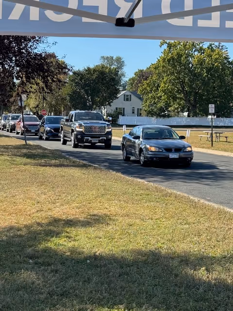 Cars drive along a paved road beside a grassy area and trees near a facility.