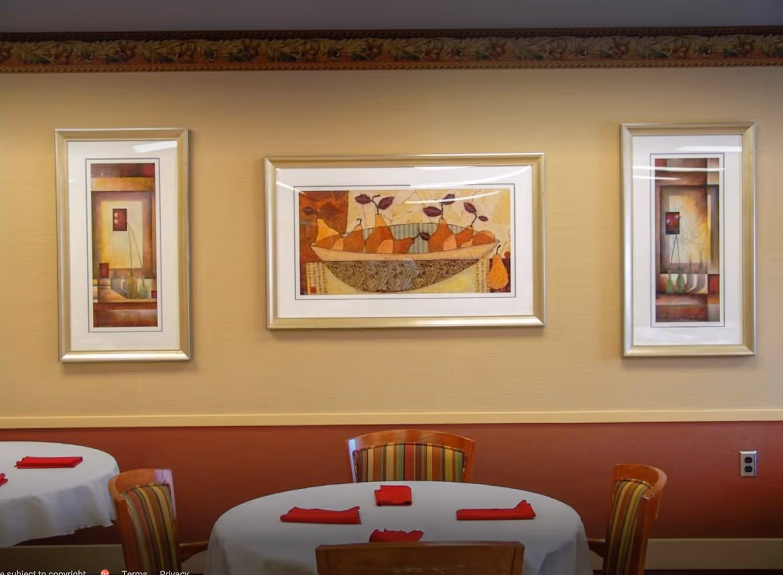 A dining room with round tables covered in white tablecloths and red napkins. The chairs have striped upholstery. On the wall, there are three framed paintings featuring abstract and still life designs with warm colors.