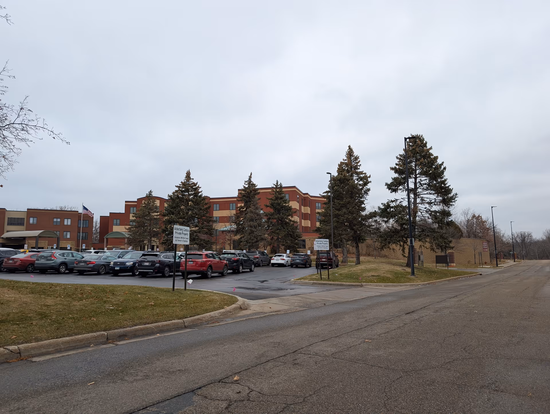 Multi-story brick assisted living building front with a parking lot, parked cars, and evergreen trees under a cloudy sky.